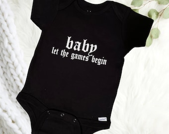 May include: Black baby onesie with white text that reads "baby let the games begin" in a gothic font.