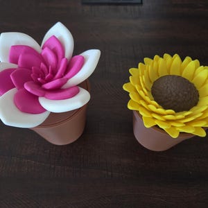 May include: Two artificial flower arrangements in small brown pots. One features a pink and white flower, the other a yellow sunflower with a brown center. The flowers are set against a dark brown wooden surface.