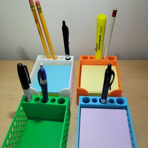 May include: Four colorful desk organizers, each with a different color: white, orange, green, and blue. Each organizer holds a stack of sticky notes and has holes for pens and pencils. Pencils, pens, and a highlighter are in the organizers.