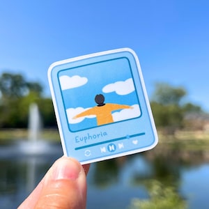 May include: A blue sticker with a cartoon illustration of a person with arms outstretched against a blue sky with white clouds. The sticker has the text "Euphoria" and music player controls.
