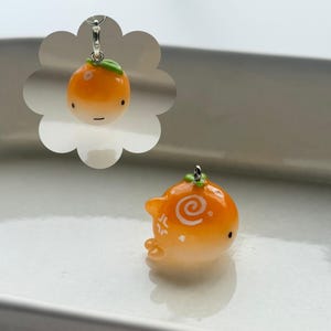 K&ouml;nnte beinhalten: Orangener Anh&auml;nger mit einem gr&uuml;nen Blatt und einer silberfarbenen Schlaufe. Der Anh&auml;nger hat ein Cartoon-Gesicht und ein Wirbeldesign. Der Anh&auml;nger befindet sich auf einer wei&szlig;en Oberfl&auml;che.