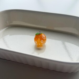 K&ouml;nnte beinhalten: Ein kleiner, orangefarbener Anh&auml;nger in Form einer Mandarine mit wei&szlig;en dekorativen Mustern. Der Anh&auml;nger hat ein kleines gr&uuml;nes Blatt und eine silberfarbene Schlaufe zum Befestigen an einer Halskette oder einem Armband. Der Anh&auml;nger liegt auf einer wei&szlig;en Oberfl&auml;che.