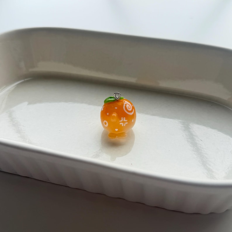 K&ouml;nnte beinhalten: Ein kleiner, orangefarbener Anh&auml;nger in Form einer Mandarine mit wei&szlig;en dekorativen Mustern. Der Anh&auml;nger hat ein kleines gr&uuml;nes Blatt und eine silberfarbene Schlaufe zum Befestigen an einer Halskette oder einem Armband. Der Anh&auml;nger liegt auf einer wei&szlig;en Oberfl&auml;che.