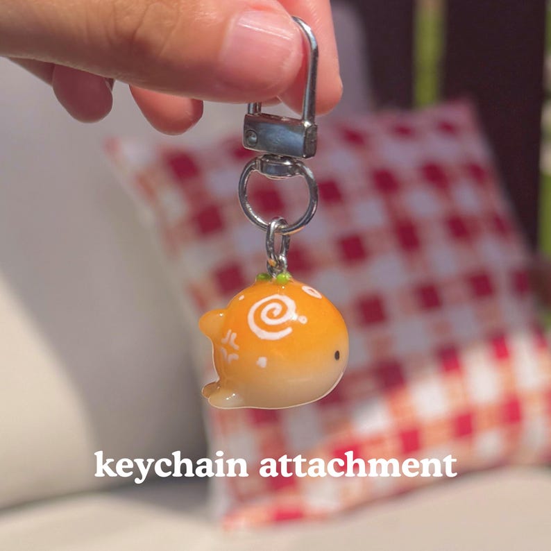 K&ouml;nnte beinhalten: Ein Schl&uuml;sselanh&auml;nger mit einer kleinen, orangefarbenen Cartoonfigur mit einem wei&szlig;en Wirbeldesign. Der Schl&uuml;sselanh&auml;nger hat einen silberfarbenen Verschluss und Ring. Der Text "keychain attachment" ist unten im Bild zu sehen.