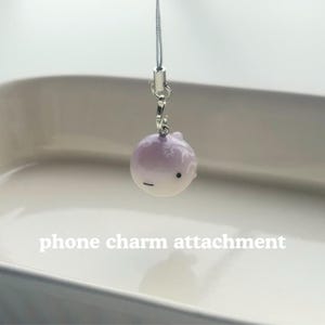 K&ouml;nnte beinhalten: Ein kleiner, runder, lila und wei&szlig;er Handy-Anh&auml;nger. Der Anh&auml;nger hat ein kleines Gesicht mit einem schwarzen Punkt und einem horizontalen Strich. Der Anh&auml;nger ist an einem silberfarbenen Metallverschluss und einer d&uuml;nnen, silbernen Kordel befestigt. Der Text "phone charm attachment" steht darunter.