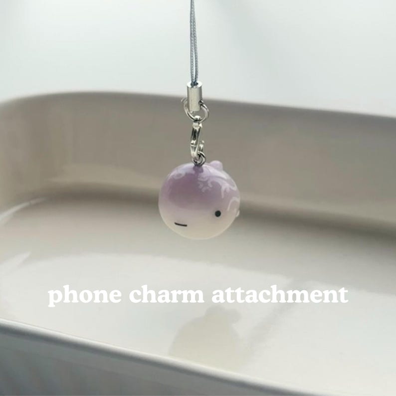 K&ouml;nnte beinhalten: Ein kleiner, runder, lila und wei&szlig;er Handy-Anh&auml;nger. Der Anh&auml;nger hat ein kleines Gesicht mit einem schwarzen Punkt und einem horizontalen Strich. Der Anh&auml;nger ist an einem silberfarbenen Metallverschluss und einer d&uuml;nnen, silbernen Kordel befestigt. Der Text "phone charm attachment" steht darunter.