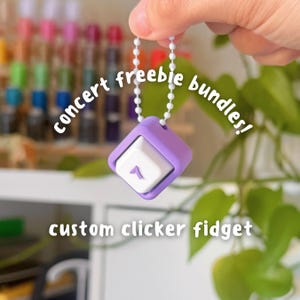 Puede incluir: Un fidget clicker personalizado en forma cuadrada con un marco morado y un botón blanco. El botón tiene un número 1 morado. El fidget está sujeto a una cadena de cuentas blancas. El texto en la imagen dice "concert freebie bundles!"