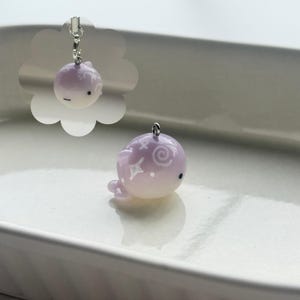 K&ouml;nnte beinhalten: Ein kleiner, lavendelfarbener Wal-Anh&auml;nger mit wei&szlig;en Stern- und Wirbelmustern. Der Anh&auml;nger hat oben eine silberfarbene Schlaufe zum Aufh&auml;ngen. Der Wal hat eine Farbverlauf von Lavendel zu Wei&szlig;. Ein niedliches Accessoire.