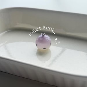 K&ouml;nnte beinhalten: Ein kleiner, runder, lavendelfarbener Anh&auml;nger mit einer silberfarbenen Schlaufe oben. Der Anh&auml;nger hat ein Cartoon-Gesicht mit einer schwarzen Linie f&uuml;r den Mund und kleinen schwarzen Punkten f&uuml;r die Augen. Der Text "meet lumi..." ist in schwarzer Schreibschrift geschrieben.
