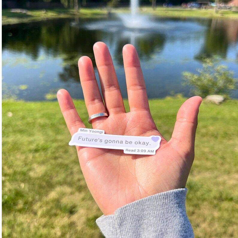 May include: A white sticker with a purple heart and the text "Min Yoongi Future's gonna be okay. Read 3:09 AM" on a hand in front of a blurry background of a pond and green grass.