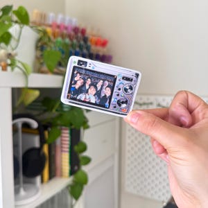 Puede incluir: Una pegatina blanca y plateada con forma de c&aacute;mara digital. La pantalla muestra un grupo de personas. La pegatina est&aacute; sujeta por una mano.
