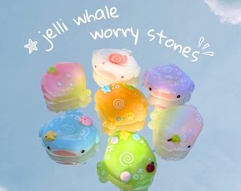 Puede incluir: Una colección de siete piedras de preocupación translúcidas con forma de ballena en varios colores pastel, incluyendo rosa, naranja, azul, verde y morado. Cada pieza presenta pequeños detalles decorativos como estrellas y espirales. Texto: jelli whale worry stones.