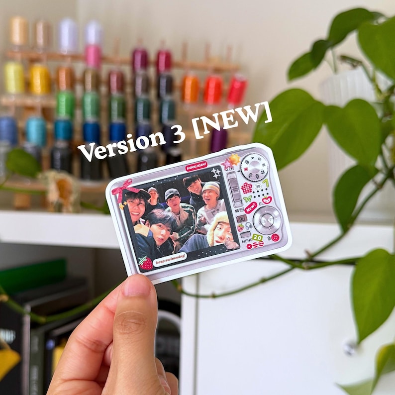 Puede incluir: Una pegatina rectangular con un dise&ntilde;o de c&aacute;mara, que presenta una foto de grupo. La pegatina tiene un borde blanco y el texto "Version 3 [NEW]" est&aacute; impreso en la parte superior. El dise&ntilde;o de la c&aacute;mara incluye botones y diales.