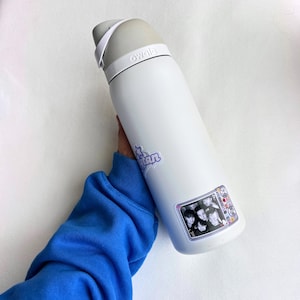 Puede incluir: Botella de agua Owala blanca con tapa y asa grises. La botella tiene dos pegatinas: una con la palabra "Titan" en morado y otra con una foto de un grupo de personas dentro de un marco de televisi&oacute;n.