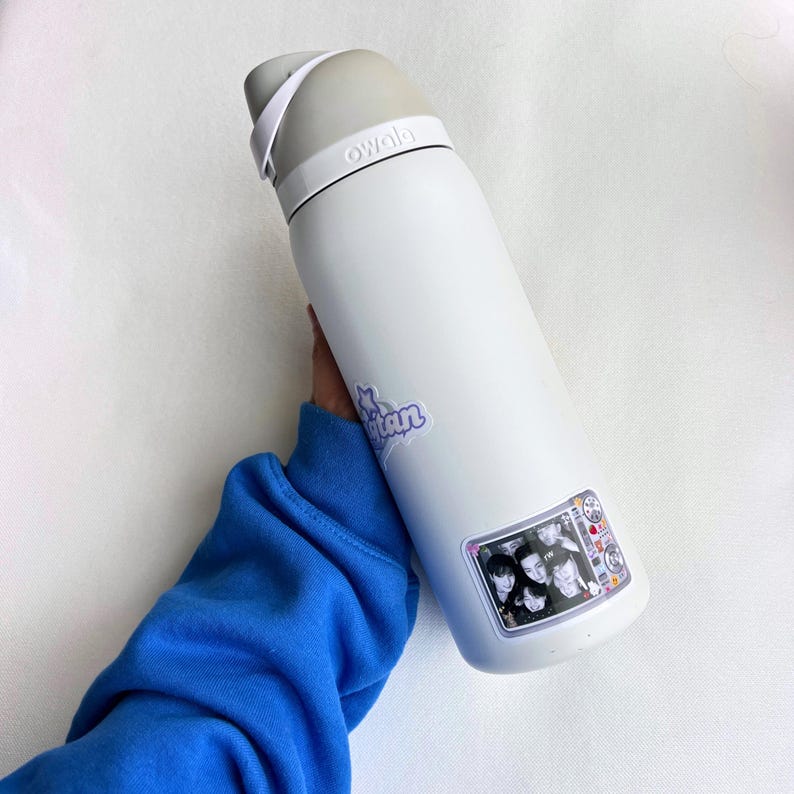 Puede incluir: Botella de agua Owala blanca con tapa y asa grises. La botella tiene dos pegatinas: una con la palabra "Titan" en morado y otra con una foto de un grupo de personas dentro de un marco de televisi&oacute;n.