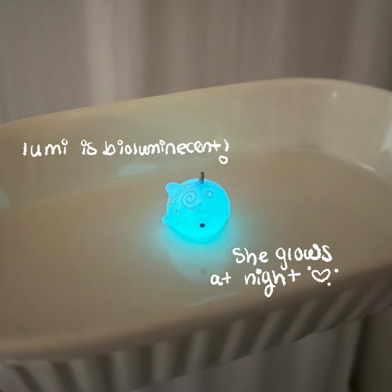 K&ouml;nnte beinhalten: Ein leuchtender, blau leuchtender Wal-Anh&auml;nger mit einer kleinen Schlaufe. Der Anh&auml;nger befindet sich auf einer wei&szlig;en Oberfl&auml;che, und der Text "lumi is bioluminescent" und "She glows at night" sind auf der Oberfl&auml;che geschrieben.