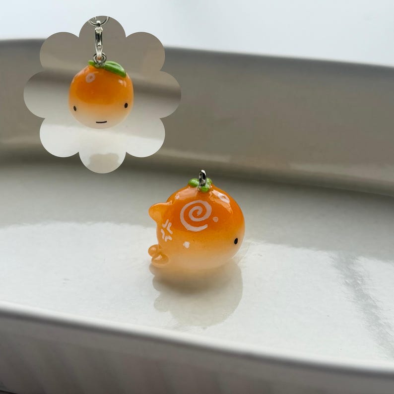 K&ouml;nnte beinhalten: Orangener Anh&auml;nger mit einem gr&uuml;nen Blatt und einer silberfarbenen Schlaufe. Der Anh&auml;nger hat ein Cartoon-Gesicht und ein Wirbeldesign. Der Anh&auml;nger befindet sich auf einer wei&szlig;en Oberfl&auml;che.