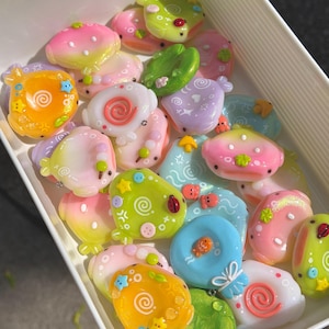 Op de afbeelding: Een verzameling kleine, kleurrijke, glanzende harsbedels in de vorm van afgeronde wezens en waterleliebladeren in een witte rechthoekige bak. De bedels hebben pastelkleurige gradi&euml;nten, wervelpatronen, kleine steraccenten en details zoals lieveheersbeestjes.