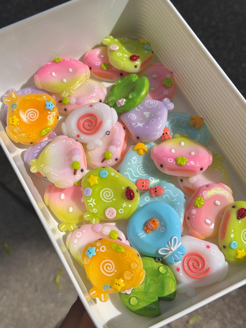Op de afbeelding: Een verzameling kleine, kleurrijke, glanzende harsbedels in de vorm van afgeronde wezens en waterleliebladeren in een witte rechthoekige bak. De bedels hebben pastelkleurige gradi&euml;nten, wervelpatronen, kleine steraccenten en details zoals lieveheersbeestjes.