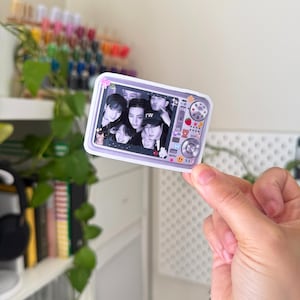 Puede incluir: Una pegatina que representa a un grupo de personas al estilo de un fotomat&oacute;n. La pegatina tiene un borde lila y un dise&ntilde;o de c&aacute;mara con elementos decorativos como osos, fresas y emoticonos. La foto muestra un grupo de personas.
