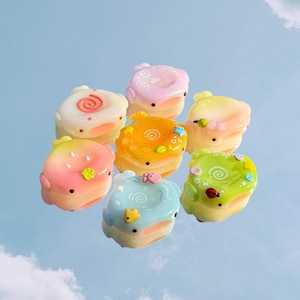 Op de afbeelding: Een verzameling van zeven kleine, afgeronde, pastelkleurige harsbedels met diverse reli&euml;fmotieven, waaronder spiralen, kleine bloemetjes, sterretjes en een lieveheersbeestje. Elk stuk heeft een gladde, glanzende afwerking en duidelijke kleurverlopen van roze en oranje tot blauw en groen.
