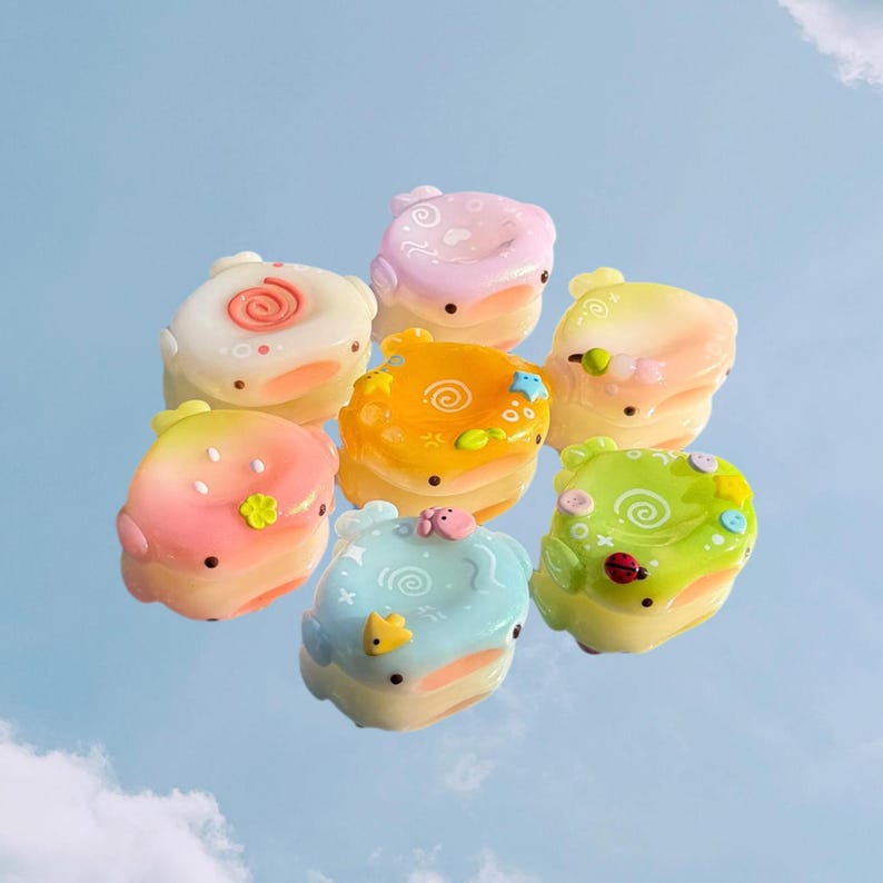Op de afbeelding: Een verzameling van zeven kleine, afgeronde, pastelkleurige harsbedels met diverse reli&euml;fmotieven, waaronder spiralen, kleine bloemetjes, sterretjes en een lieveheersbeestje. Elk stuk heeft een gladde, glanzende afwerking en duidelijke kleurverlopen van roze en oranje tot blauw en groen.