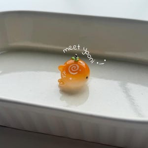 K&ouml;nnte beinhalten: Ein kleiner, orangefarbener Wal-Anh&auml;nger mit einem Spiralmuster und dem Text "meet yooni". Der Anh&auml;nger hat einen gr&uuml;nen Akzent oben und kleine wei&szlig;e Details. Er ist vor einem wei&szlig;en Hintergrund platziert.