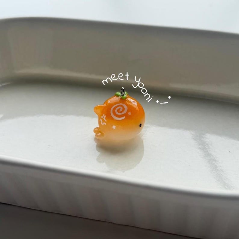 K&ouml;nnte beinhalten: Ein kleiner, orangefarbener Wal-Anh&auml;nger mit einem Spiralmuster und dem Text "meet yooni". Der Anh&auml;nger hat einen gr&uuml;nen Akzent oben und kleine wei&szlig;e Details. Er ist vor einem wei&szlig;en Hintergrund platziert.
