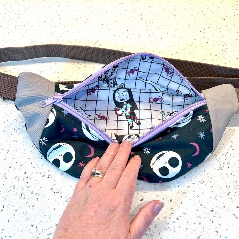 May include: A black and green fanny pack with a zipper closure. The fanny pack has a black and white skull pattern and a purple lining. The lining features a repeated pattern of a girl with a spider web and a spider.