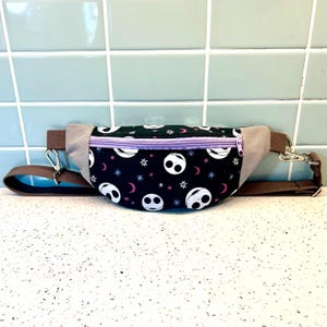 May include: A black fanny pack with a purple zipper and a repeating pattern of white Jack Skellington faces on a black background with pink and white stars and crescent moons. The fanny pack has a brown adjustable strap.