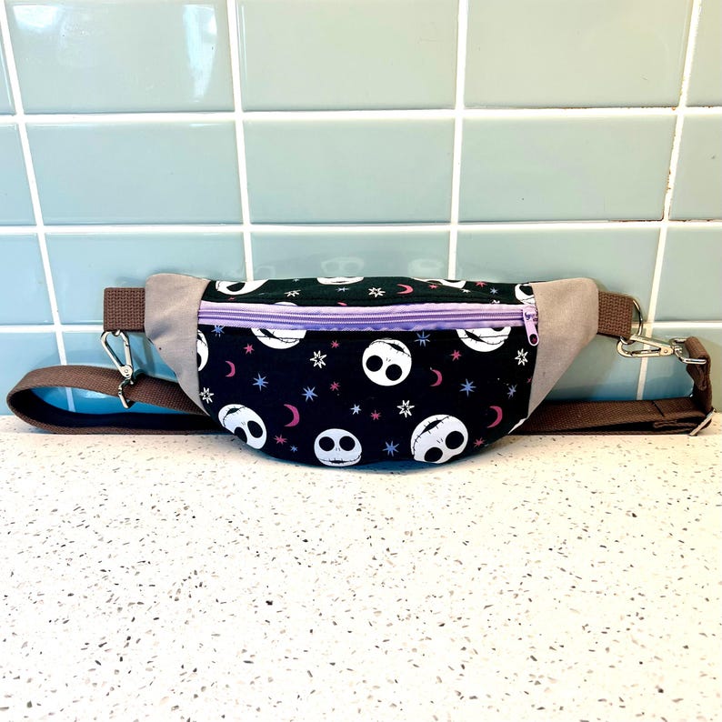 May include: A black fanny pack with a purple zipper and a repeating pattern of white Jack Skellington faces on a black background with pink and white stars and crescent moons. The fanny pack has a brown adjustable strap.