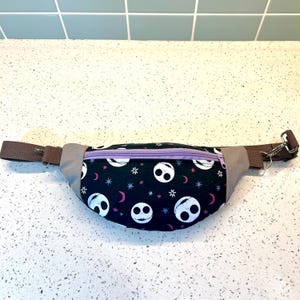 May include: A black fanny pack with a purple zipper and a repeating pattern of white Jack Skellington faces on a black background with stars and crescent moons.