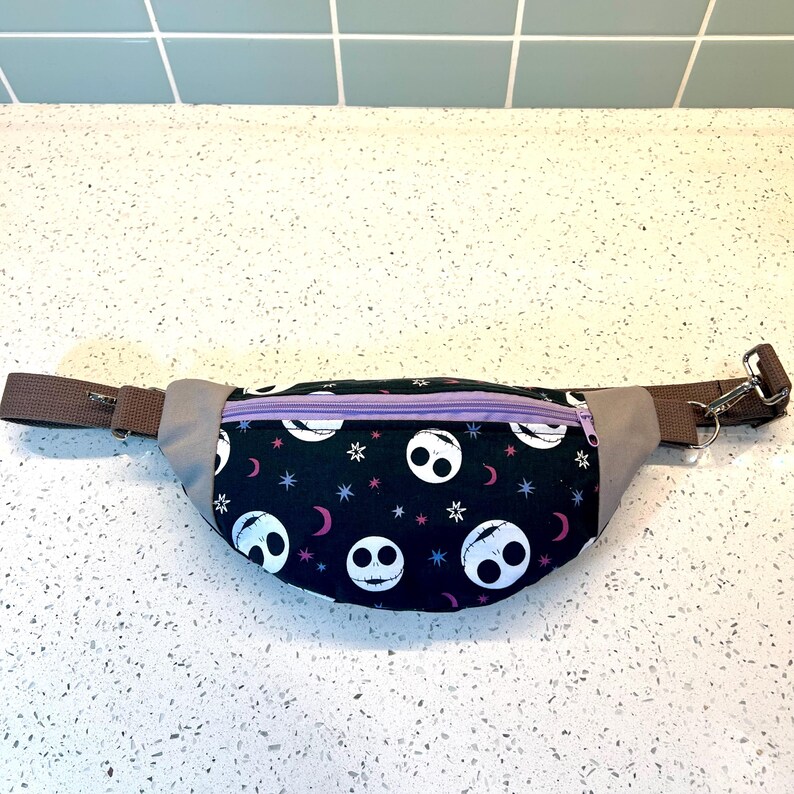 May include: A black fanny pack with a purple zipper and a repeating pattern of white Jack Skellington faces on a black background with stars and crescent moons.