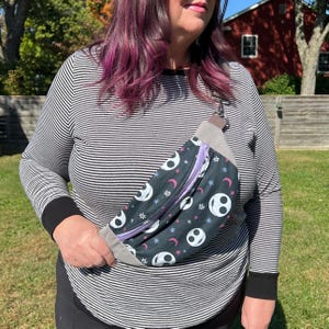 May include: A black and white fanny pack with a purple zipper and a repeating pattern of Jack Skellington's face and stars. The fanny pack is worn across the body of a person wearing a striped shirt.