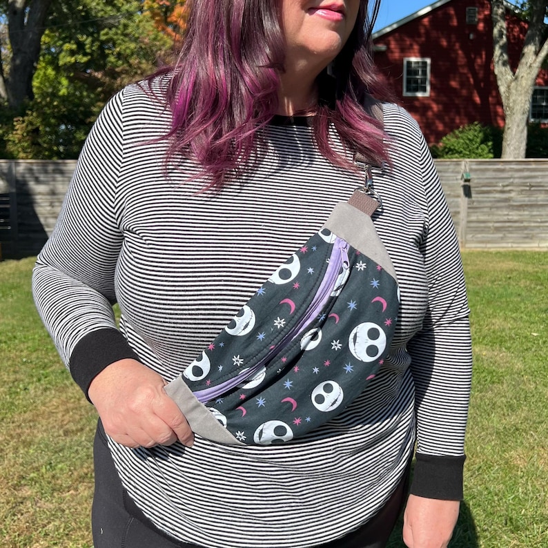 May include: A black and white fanny pack with a purple zipper and a repeating pattern of Jack Skellington's face and stars. The fanny pack is worn across the body of a person wearing a striped shirt.