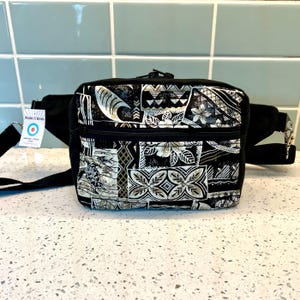 May include: Black fanny pack with a zipper closure and a white and gold patterned design. The pattern features Hawaiian-inspired motifs, including flowers, waves, and geometric shapes. The fanny pack has a black adjustable strap with a silver buckle. The tag attached to the strap reads "Made in Hawaii".