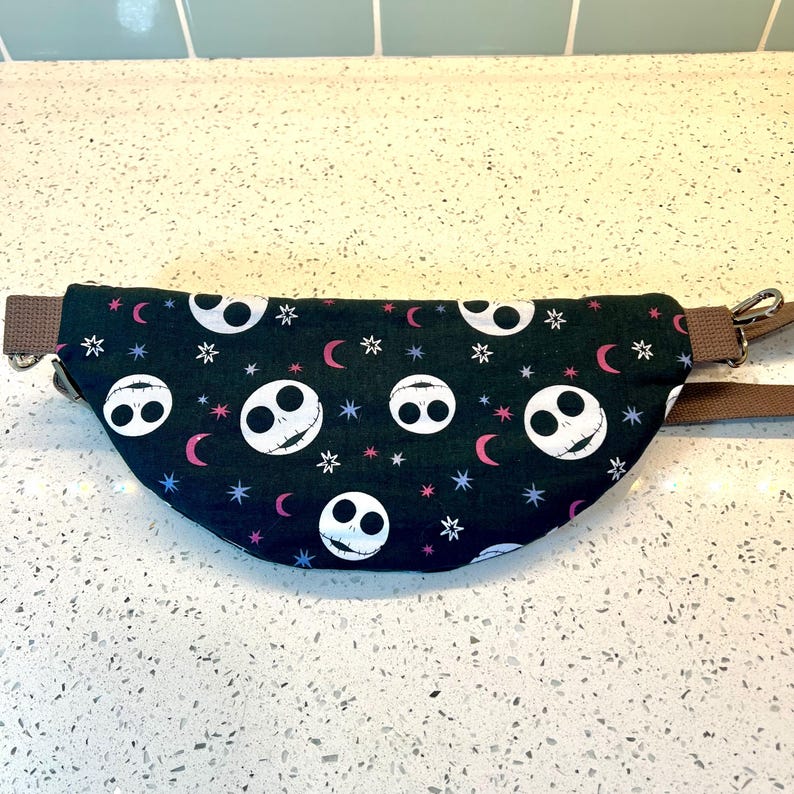 May include: A black fanny pack with a repeating pattern of white Jack Skellington faces, pink crescent moons, and white stars on a dark green background. The fanny pack has a brown adjustable strap.