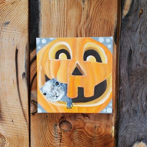 Può includere: Una tela dipinta a mano che presenta una zucca arancione sorridente con un piccolo scoiattolo grigio che spunta dall'interno. La zucca ha uno stelo nero e una faccia di zucca nera.