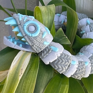May include: A gray and blue segmented snake toy with a textured surface and a toothy mouth. The toy is posed on a bed of green leaves.