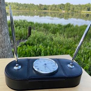 May include: A black leather desk organizer with a silver clock and two silver pen holders. The clock face has Roman numerals and the hands are pointing to 10:10.