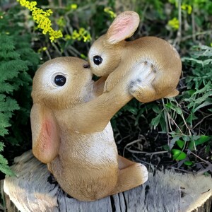 May include: A brown bunny statue holding a smaller bunny. The statue has large, dark eyes and is set against a backdrop of greenery. The bunnies are positioned on a tree stump, creating a charming outdoor scene.