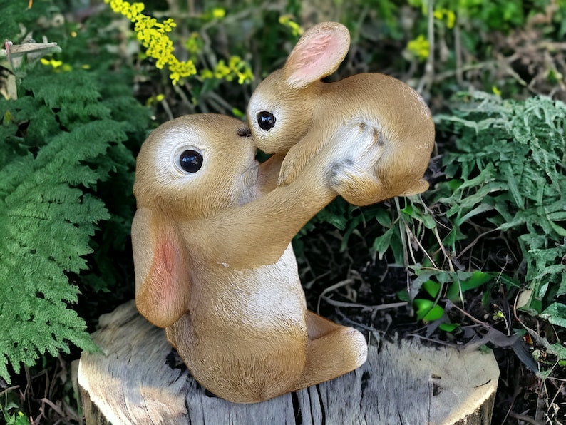 May include: A brown bunny statue holding a smaller bunny. The statue has large, dark eyes and is set against a backdrop of greenery. The bunnies are positioned on a tree stump, creating a charming outdoor scene.