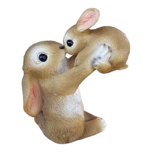 May include: A brown bunny figurine holding a smaller bunny. The larger bunny is lifting the smaller bunny up, with their noses touching. The figurine has large, dark eyes and pink accents on the ears.