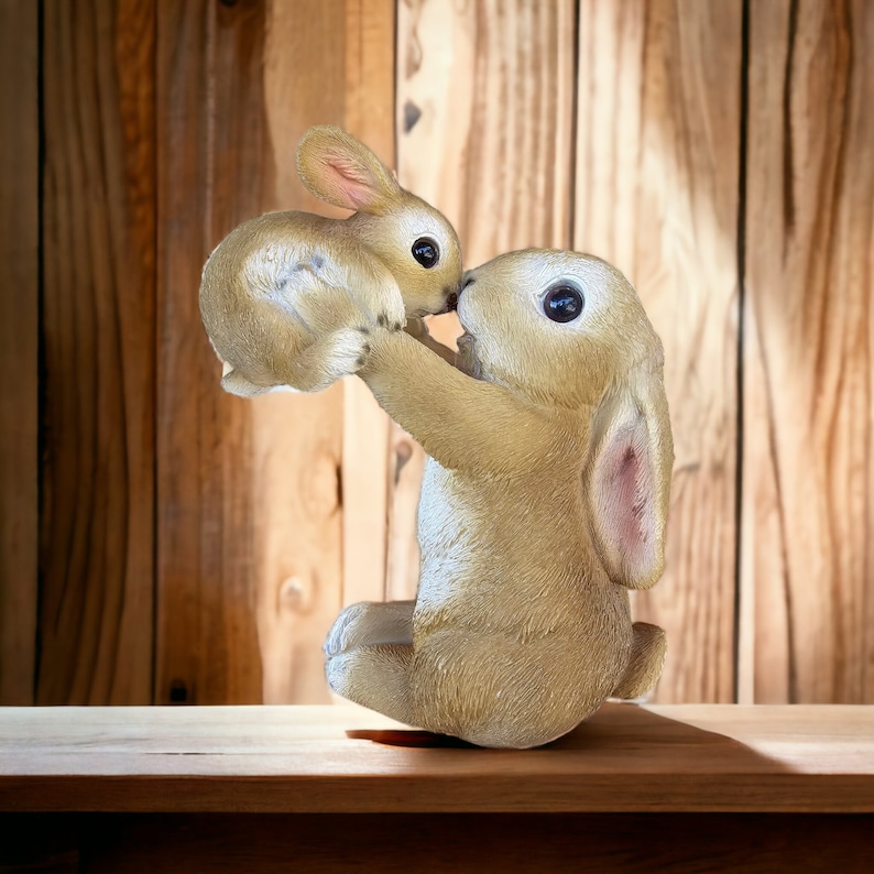 May include: A light brown bunny figurine holding a smaller bunny. The bunnies have large black eyes and pink-lined ears. The figurine is set against a wooden background.