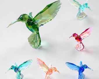 May include: A collection of six faceted crystal hummingbird figurines in various colors including green, light blue, pink, teal, orange, and dark blue. Each bird is depicted in a mid-flight pose with wings extended and pointed beaks.