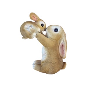 May include: A light brown bunny figurine holding a smaller bunny. The larger bunny is sitting with its legs tucked under, holding the smaller bunny up to its face. The bunnies have large black eyes and pink inner ears.