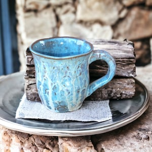 May include: A blue ceramic mug with a white glaze and a speckled pattern. The mug is sitting on a black plate and a piece of hessian fabric.