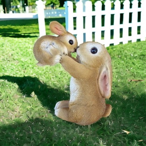 May include: A brown bunny statue holding a smaller bunny. The statues are on a green lawn with a white picket fence in the background. The bunnies have large, dark eyes and long ears. A charming garden decoration.
