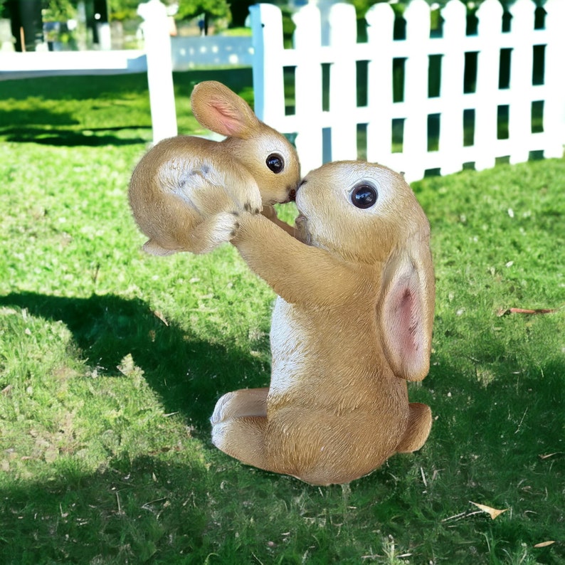 May include: A brown bunny statue holding a smaller bunny. The statues are on a green lawn with a white picket fence in the background. The bunnies have large, dark eyes and long ears. A charming garden decoration.