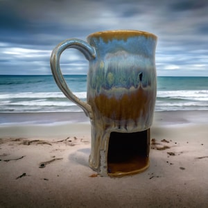 May include: A ceramic mug with a blue, brown, and white glaze. The mug has a unique design with a cutout on the bottom. The mug is sitting on a sandy beach.